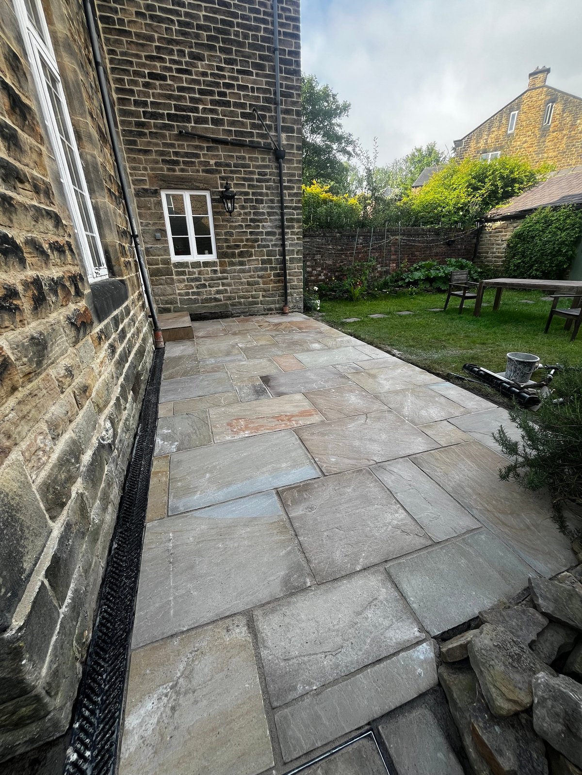 Stone patio extending from brick cottage with garden featuring green lawn, wooden bench, and hedged fence in background