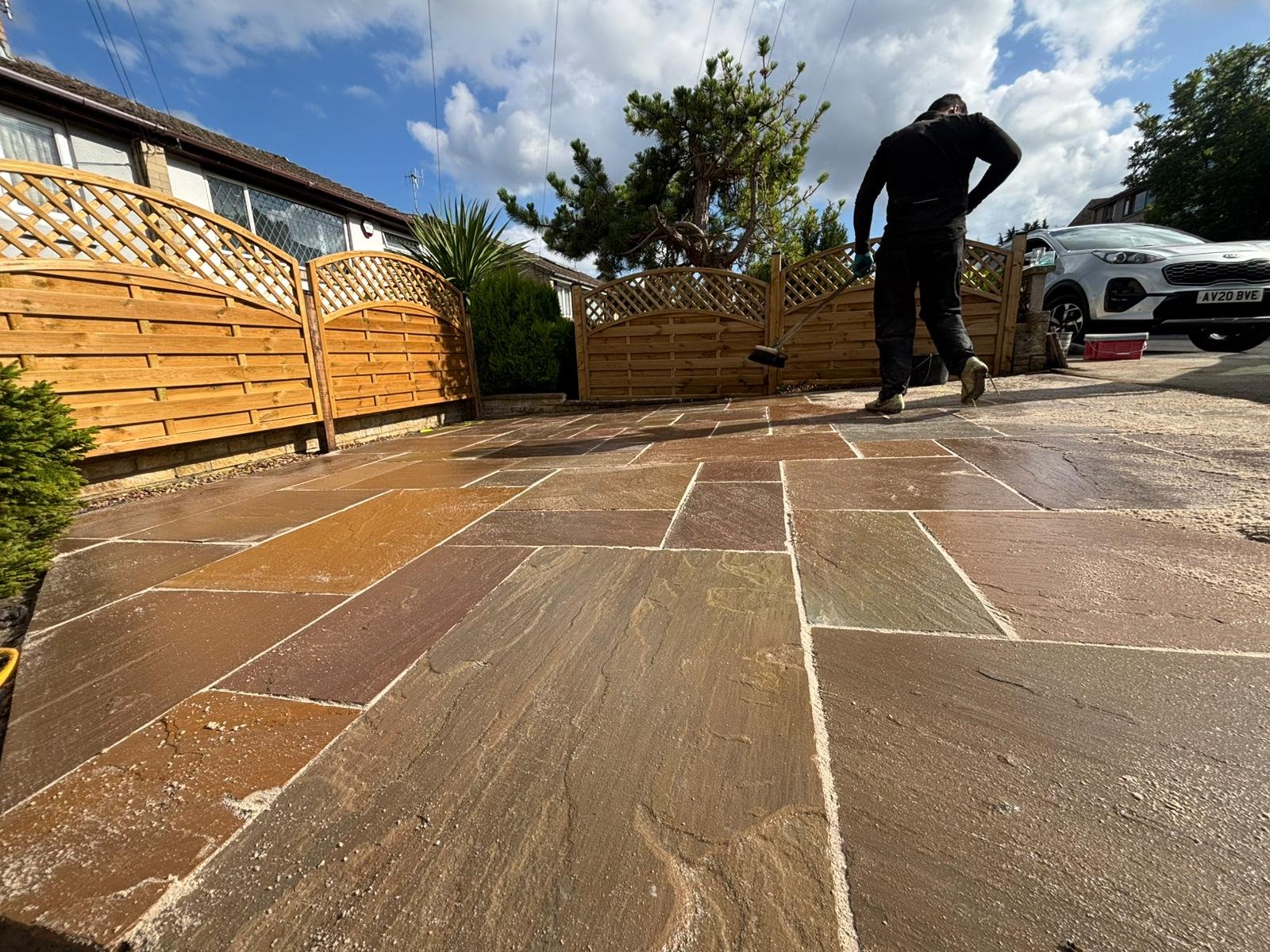 Person in black clothing sweeping a wet residential driveway with geometric paving patterns, bordered by wooden fences and parked vehicles